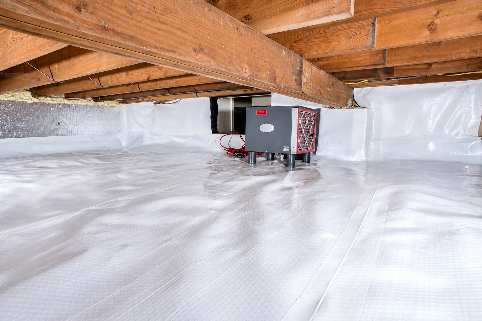 A sealed crawl space with white vapor barrier across the floor and walls and a compact testing machine beneath exposed joists.