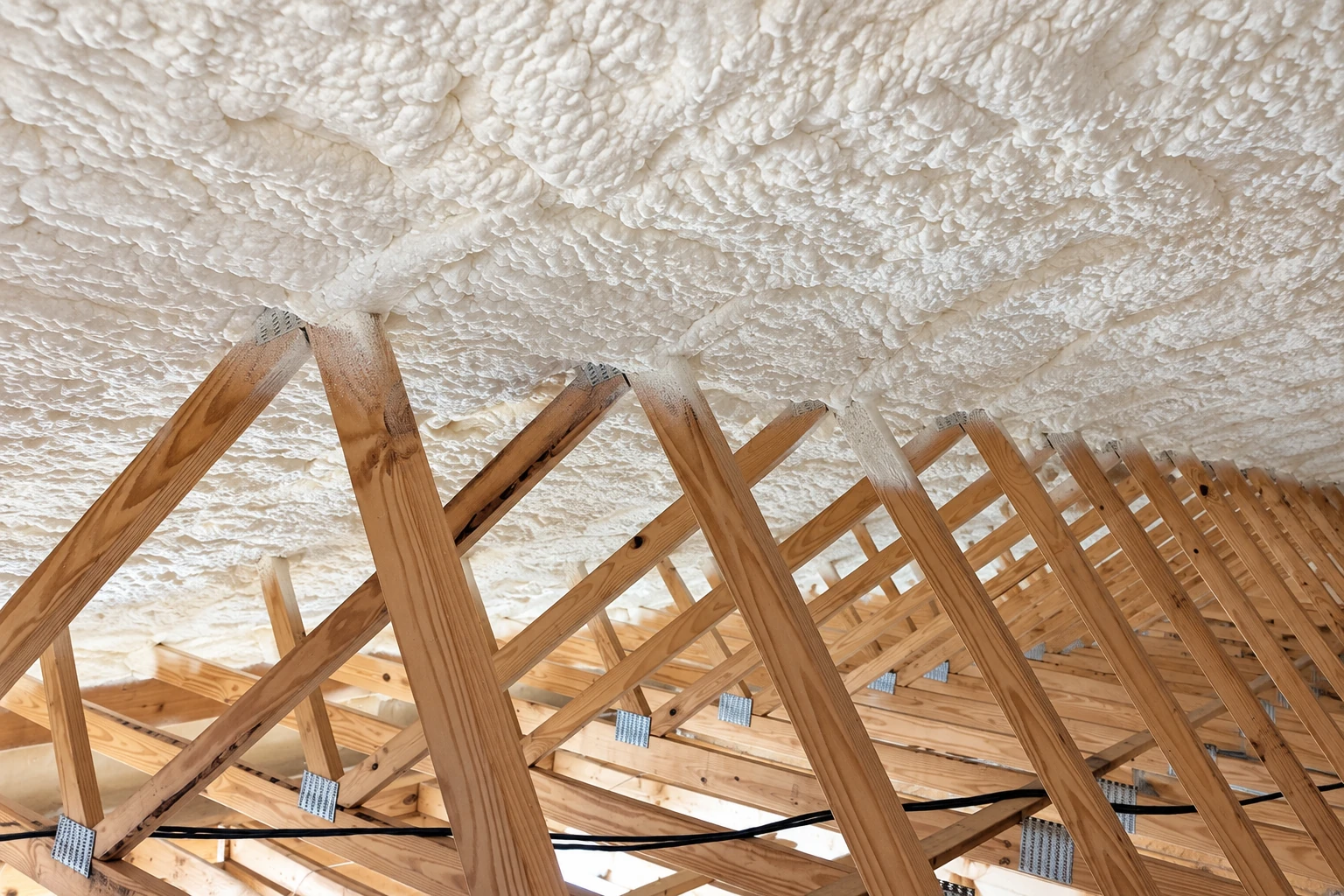 Wooden roof trusses beneath a continuous, textured layer of white spray foam insulation.