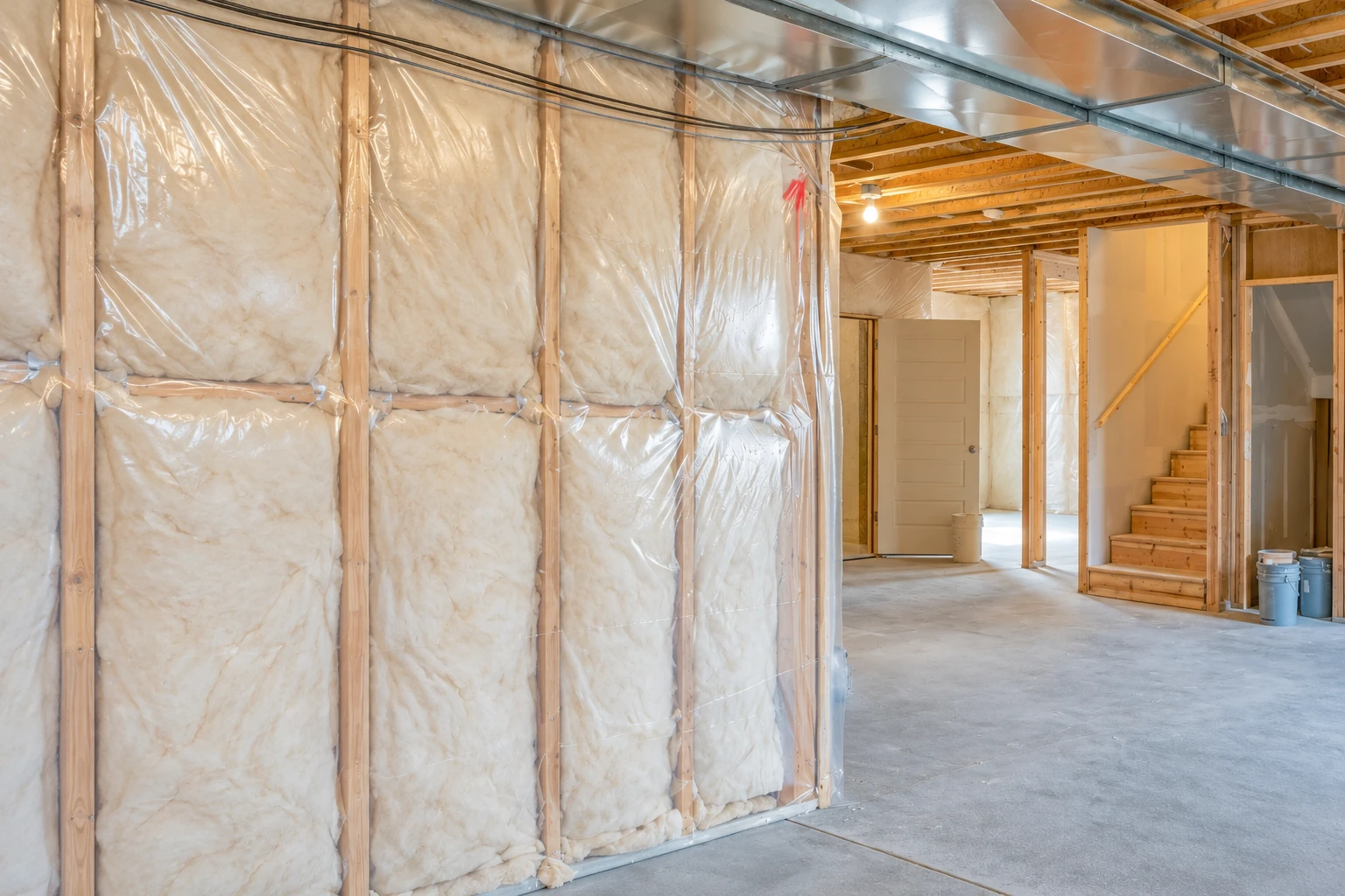 Fiberglass batts fill plastic-covered wall framing in an unfinished basement with exposed ceiling joists and stairs.