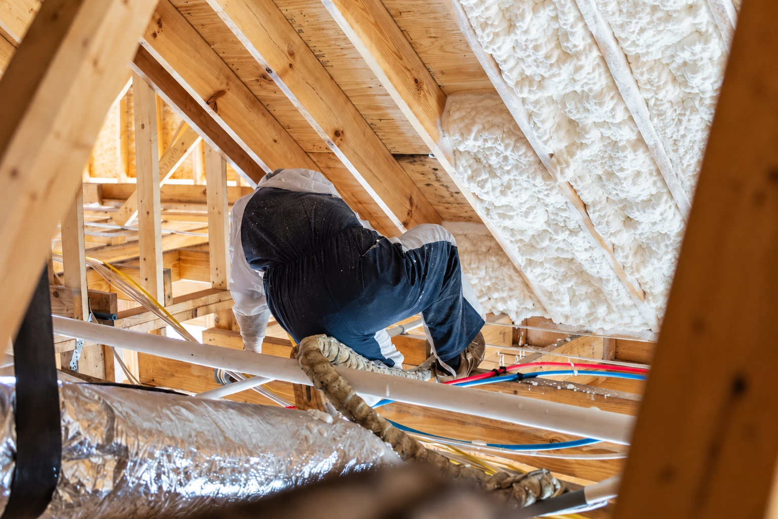 Worker in protective clothing applies white foam insulation between exposed attic rafters amid timber framing and pipes.
