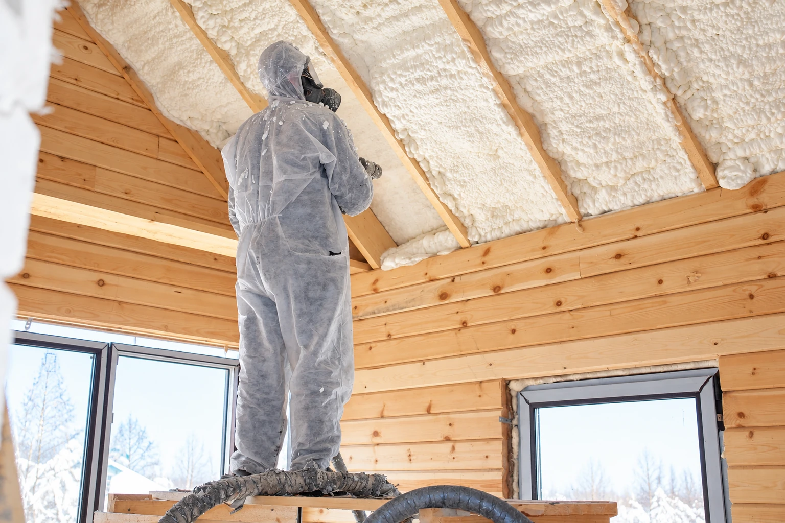 A worker in protective gear sprays white foam insulation between the exposed rafters of a wooden room.