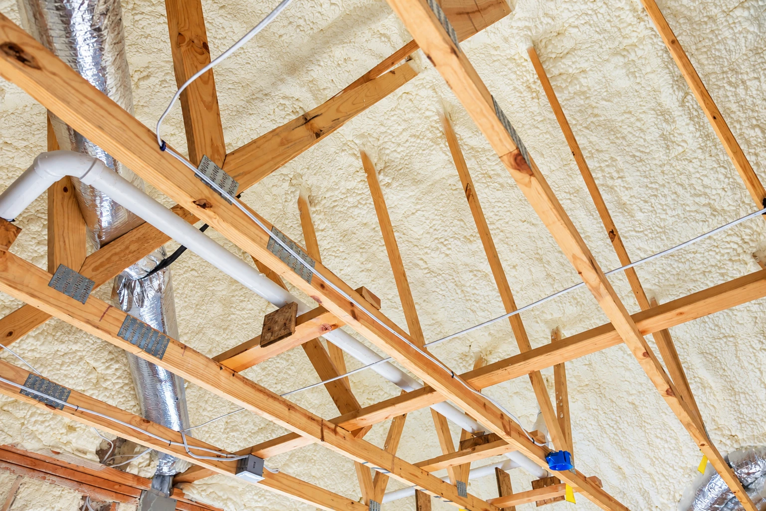 Spray foam insulation covers the roof underside between exposed wooden trusses, with pipes and wiring crossing the attic.