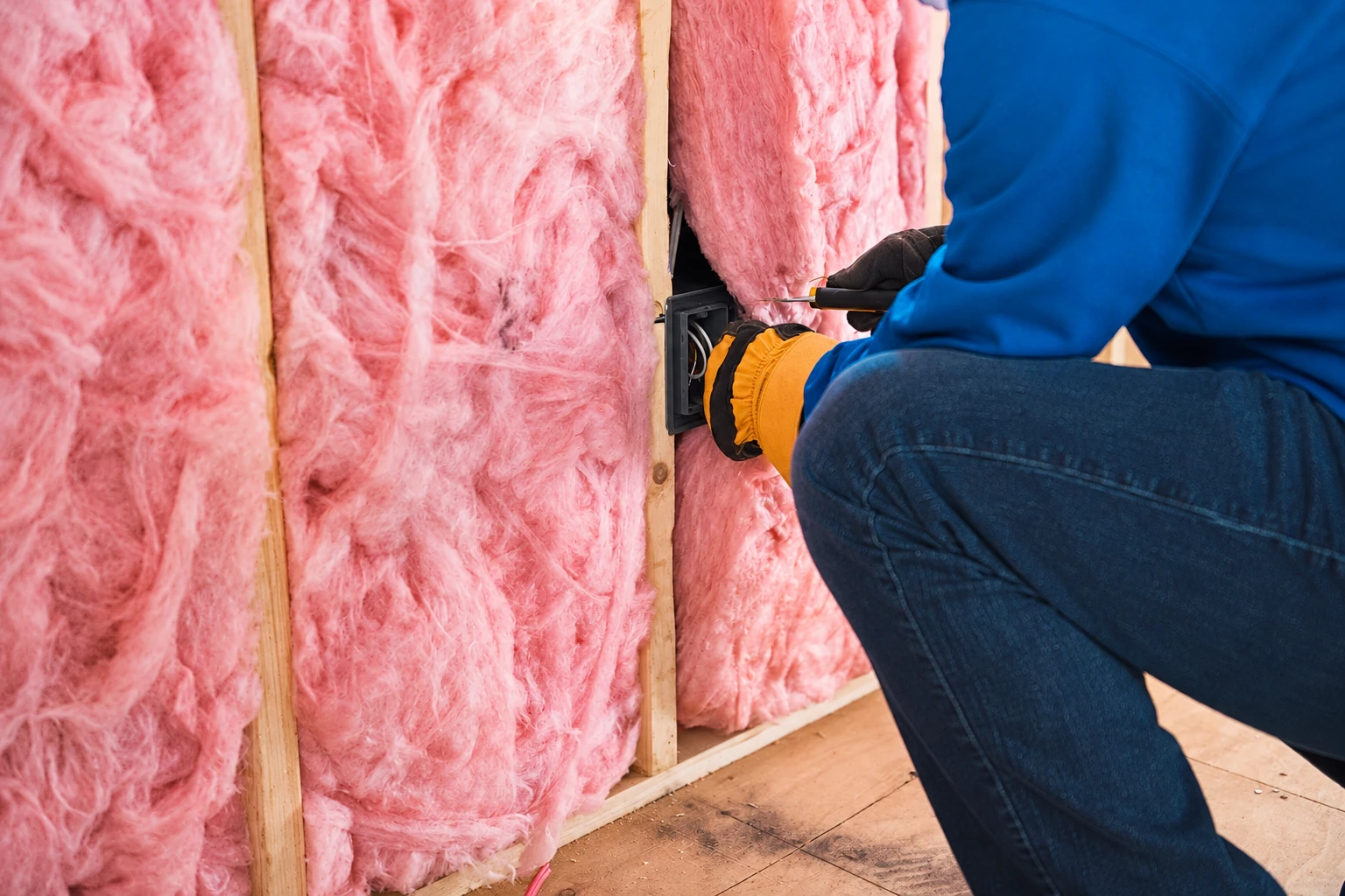 Pink fiberglass batts fill wall stud bays as a worker in blue clothing adjusts an electrical outlet.